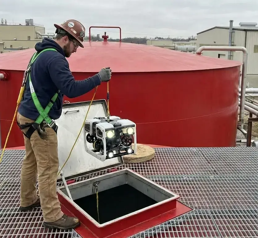 Operator w kasku i uprzęży zabezpieczającej opuszcza drona inspekcyjnego przez otwarty właz dachowy do wnętrza zbiornika przemysłowego.